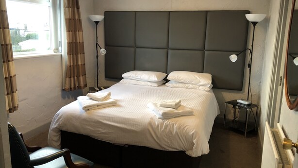 A neatly made bed with white linens and towels, a headboard, bedside lamps, a chair, and a small table with a device.