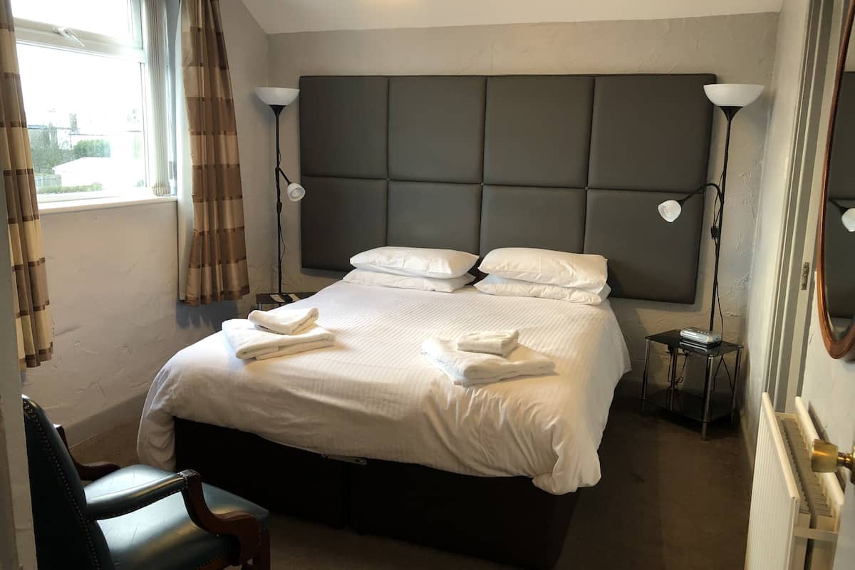 Cama bem arrumada com lençóis e toalhas brancos, cabeceira, abajures, uma cadeira e uma pequena mesa com um dispositivo.