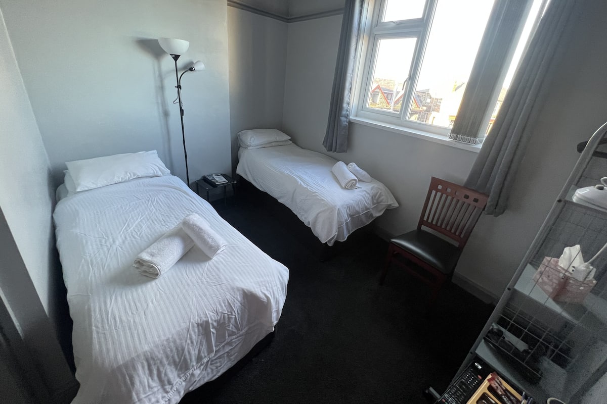 Quarto de hotel com duas camas de solteiro, uma escrivaninha com abajur, uma cadeira e uma janela com cortinas.