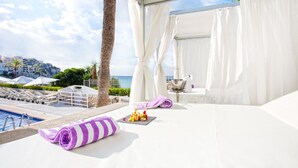 Un balcón soleado con piscina, cortinas blancas y una toalla rayada en morado y blanco.