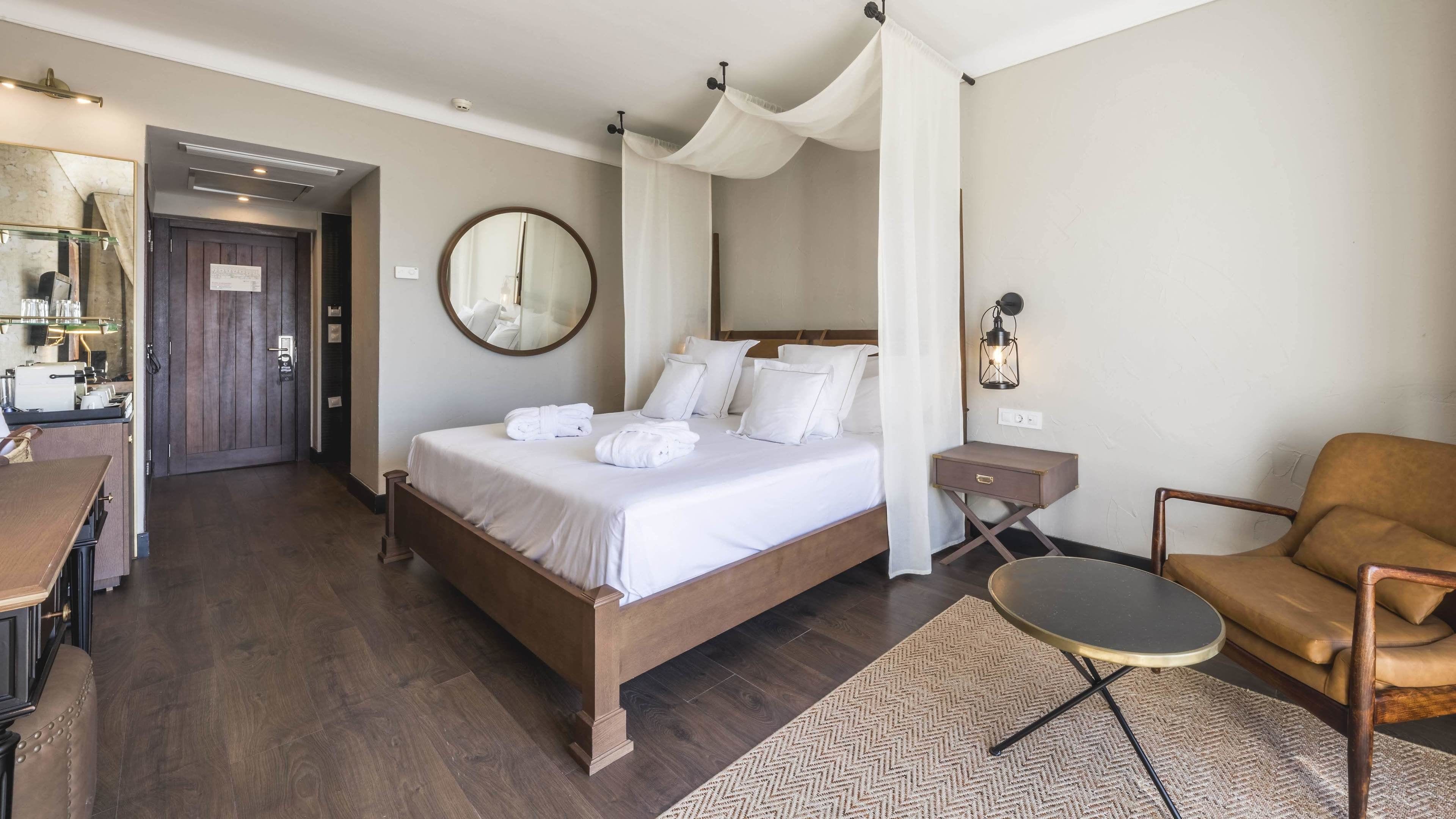 A hotel room with a large bed, a bedside table, a chair, a small table, and a mirror.