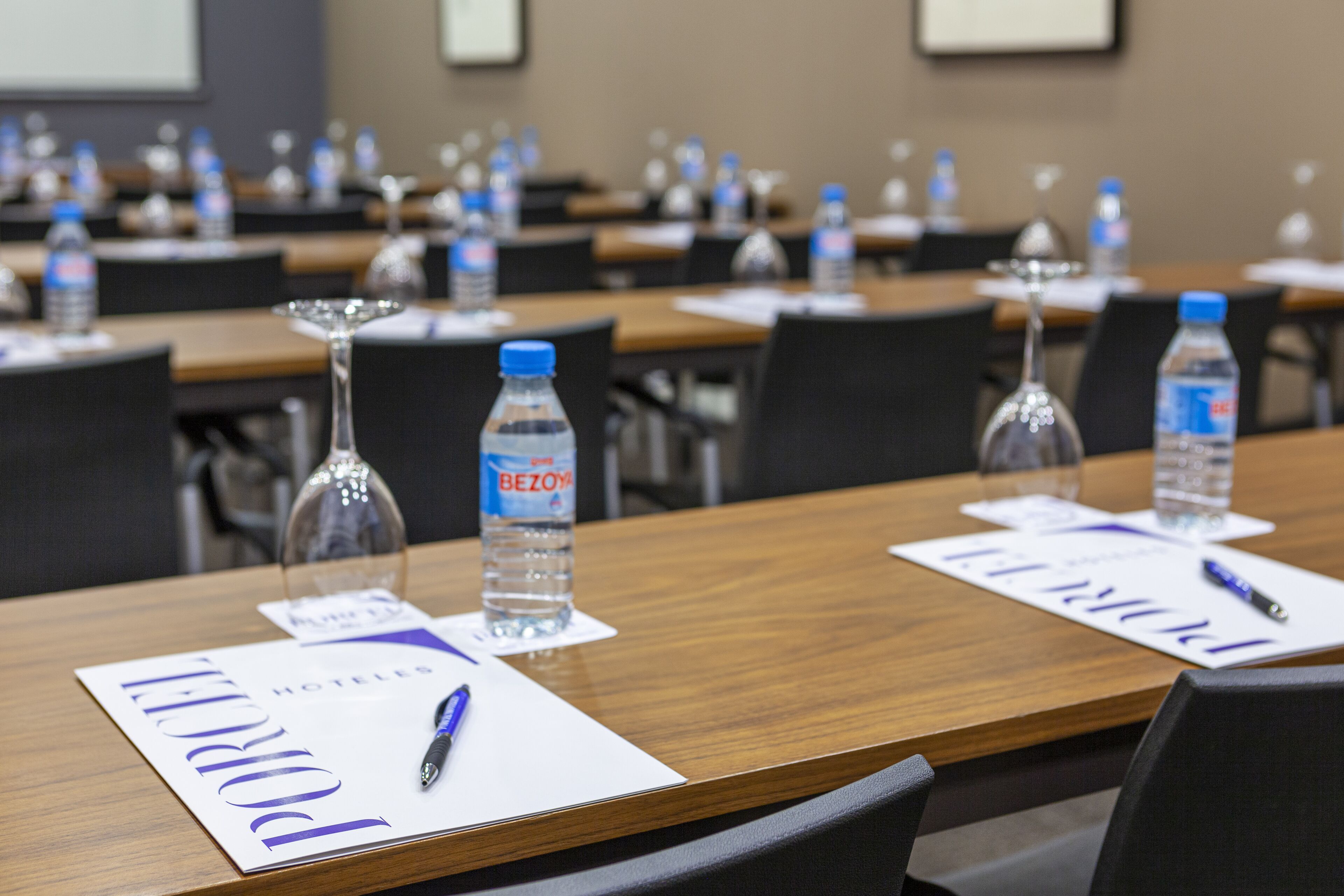 Un salón de conferencias con mesas dispuestas para reuniones, cada una con una botella de agua, un vaso y un bloc de notas.