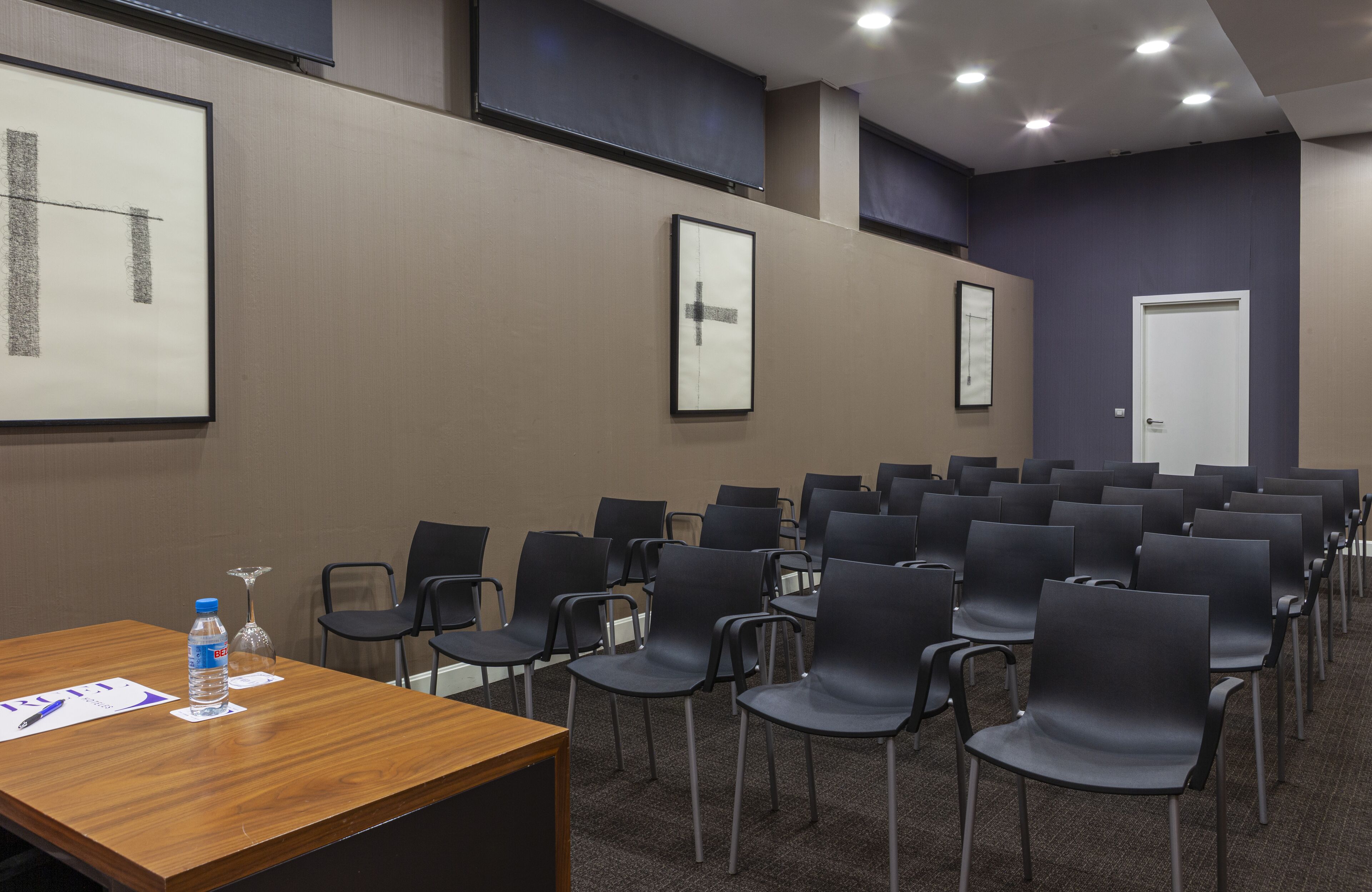 Un salón de conferencias con filas de sillas negras, una mesa de madera con una botella de agua y documentos, y obras de arte enmarcadas en las paredes.