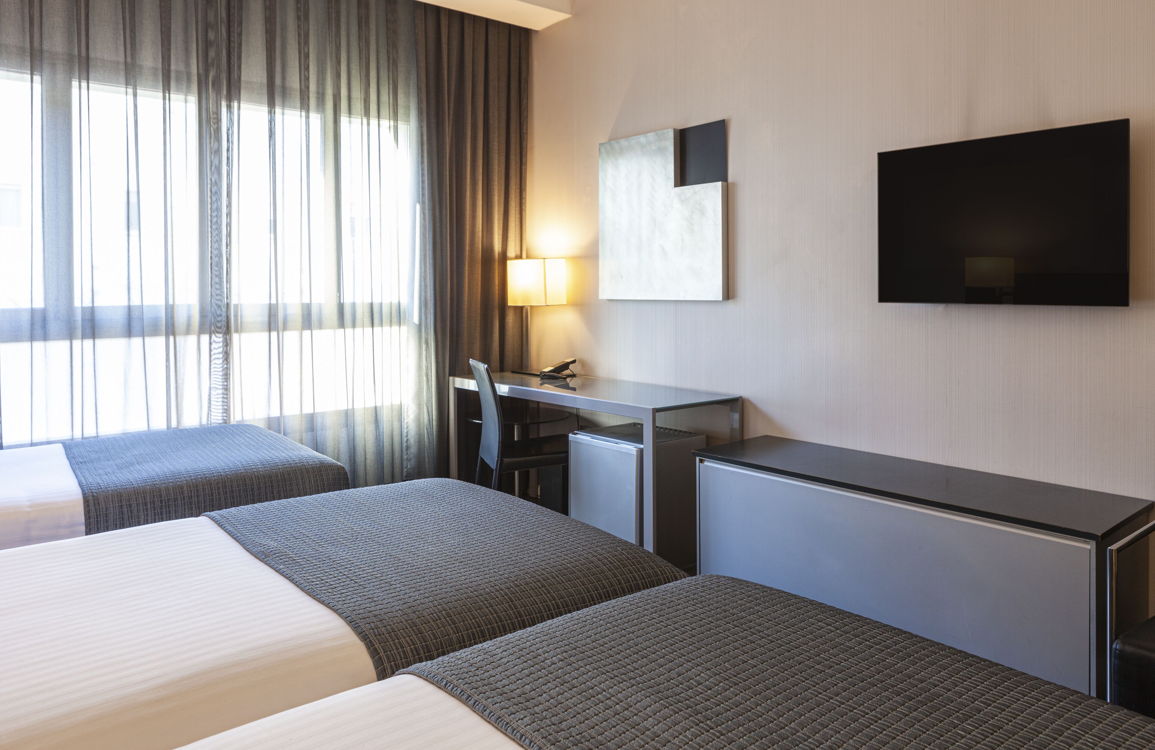 Habitació d'hotel amb dos llits, un escriptori, una cadira, un televisor i una finestra amb cortines.