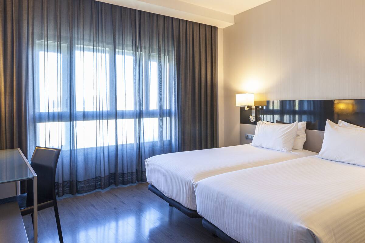 A hotel room with two beds, a desk, and a large window with curtains.