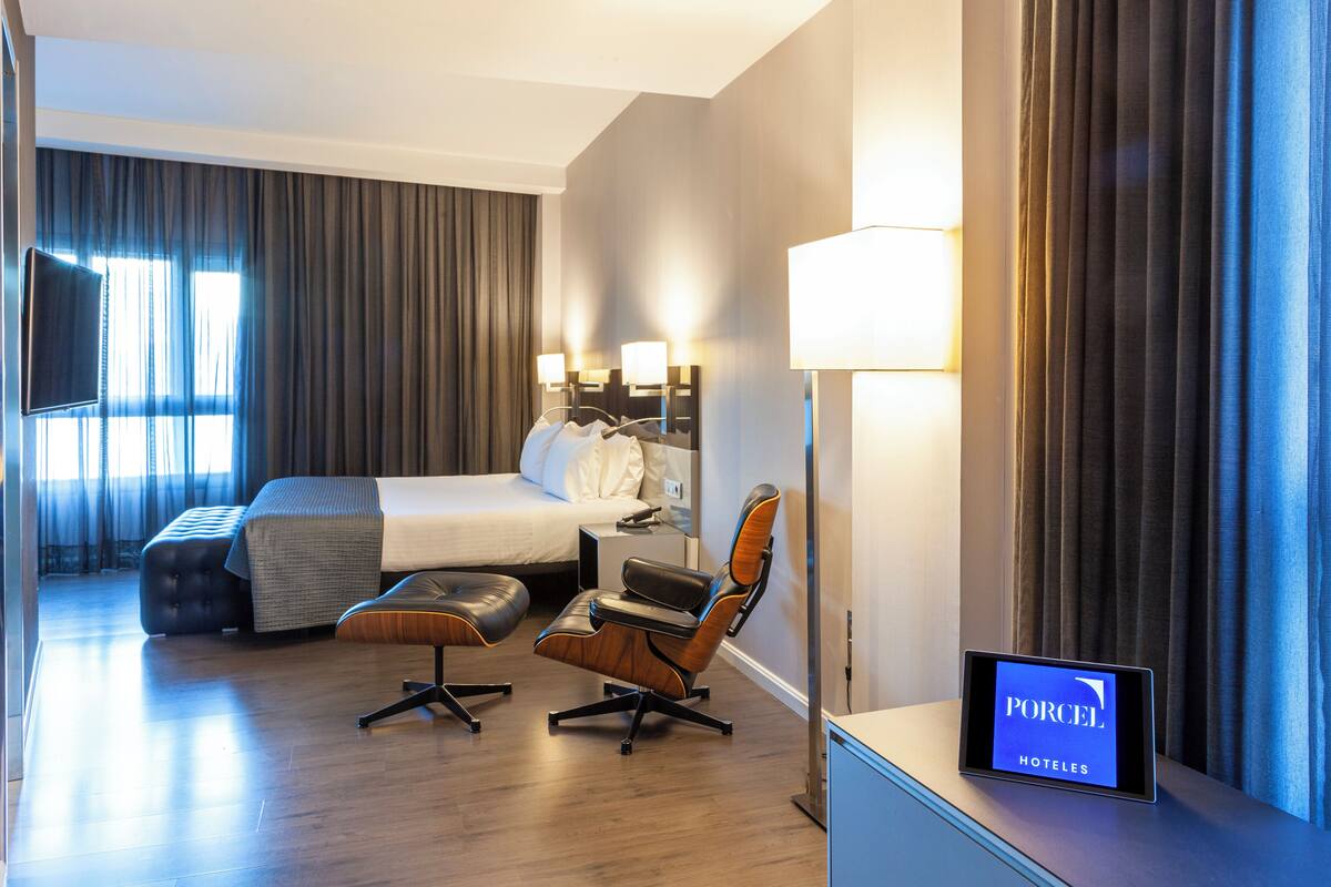 A modern hotel room with a bed, two chairs, a desk, and a TV.