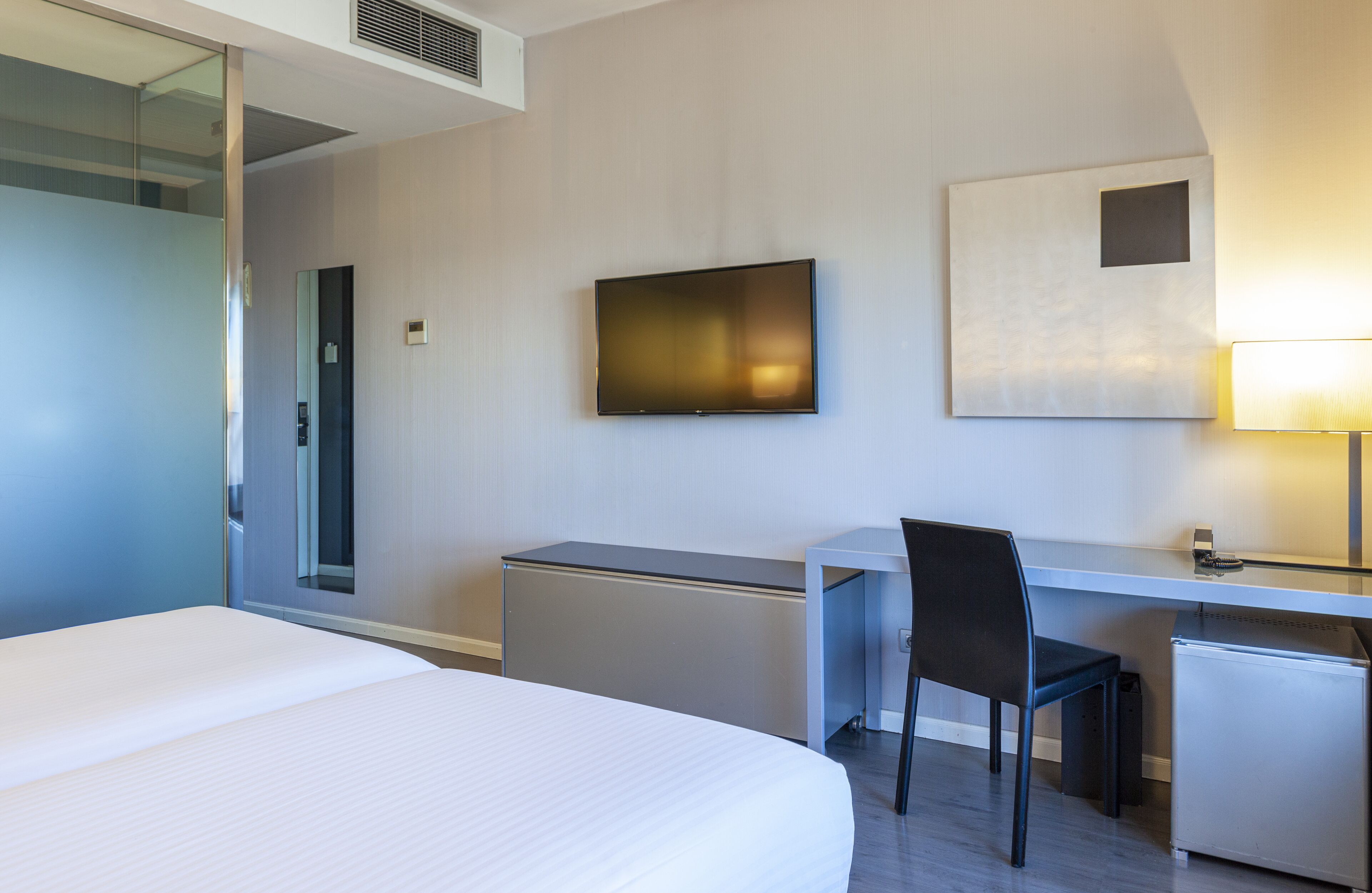 Habitación de hotel con cama, escritorio, silla, televisión, refrigerador pequeño y un cuadro colgado en la pared.