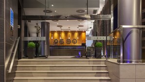 A modern hotel lobby with a staircase leading to the reception area, featuring a wooden wall with circular decorations and a glass partition. - Hotel Porcel Torneo (Seville)