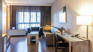 Quarto de hotel moderno com sofá, mesa de centro de vidro, escrivaninha com luminária e vista para a cidade através de grandes janelas.