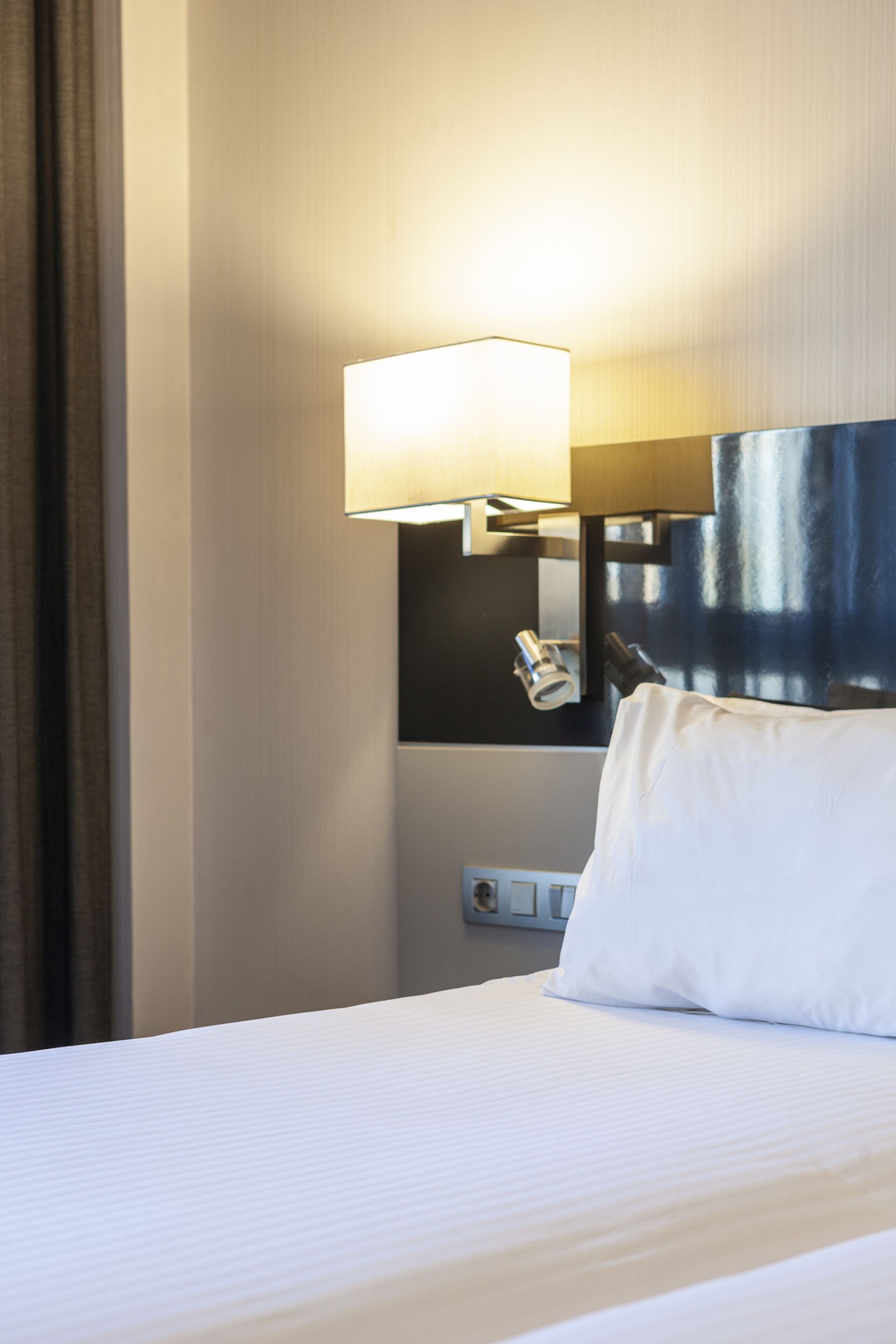 A hotel room with a neatly made bed, a bedside lamp, and a wall-mounted telephone.