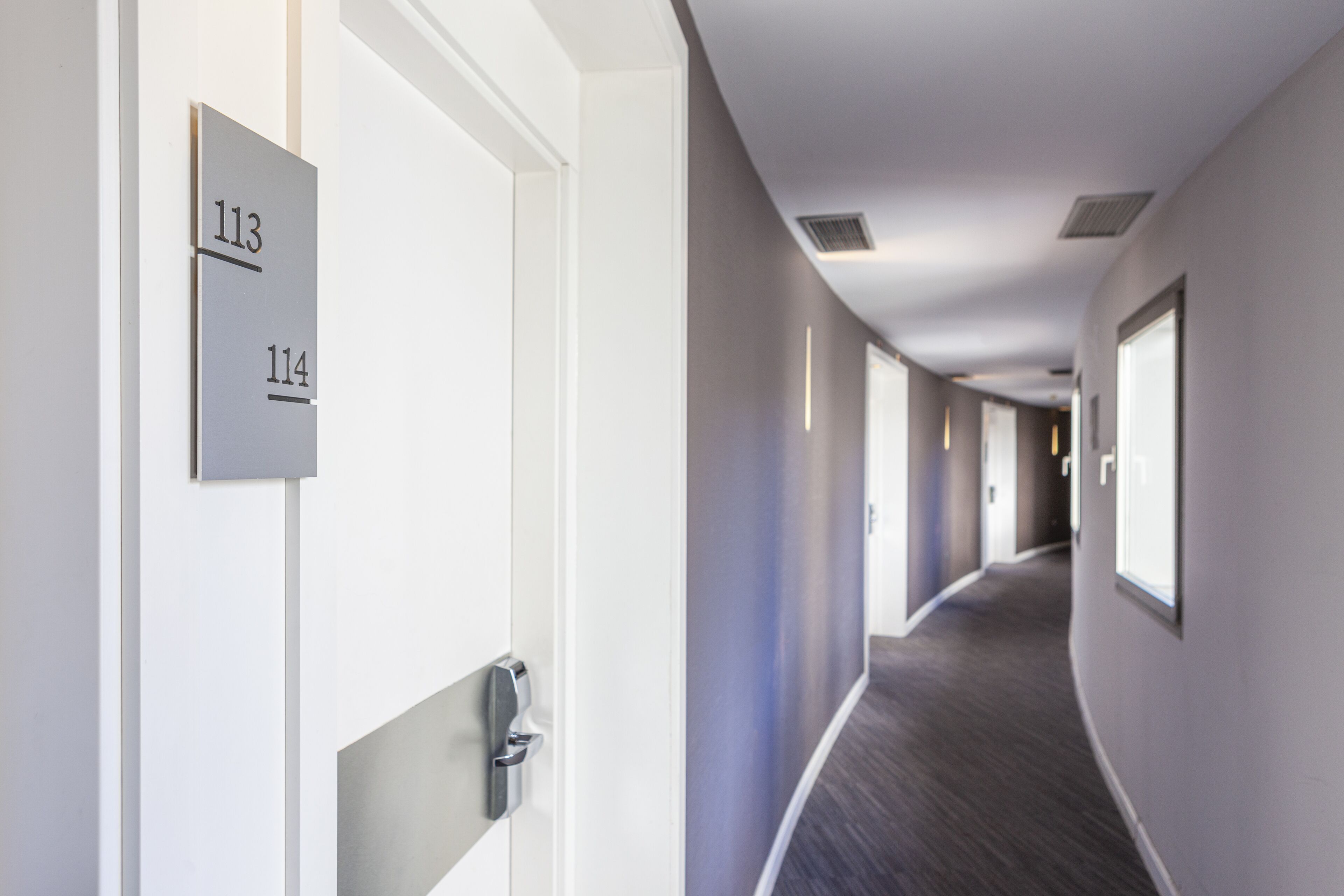 A modern hotel corridor with doors numbered 113 and 114, featuring a clean, minimalist design and a carpeted floor.