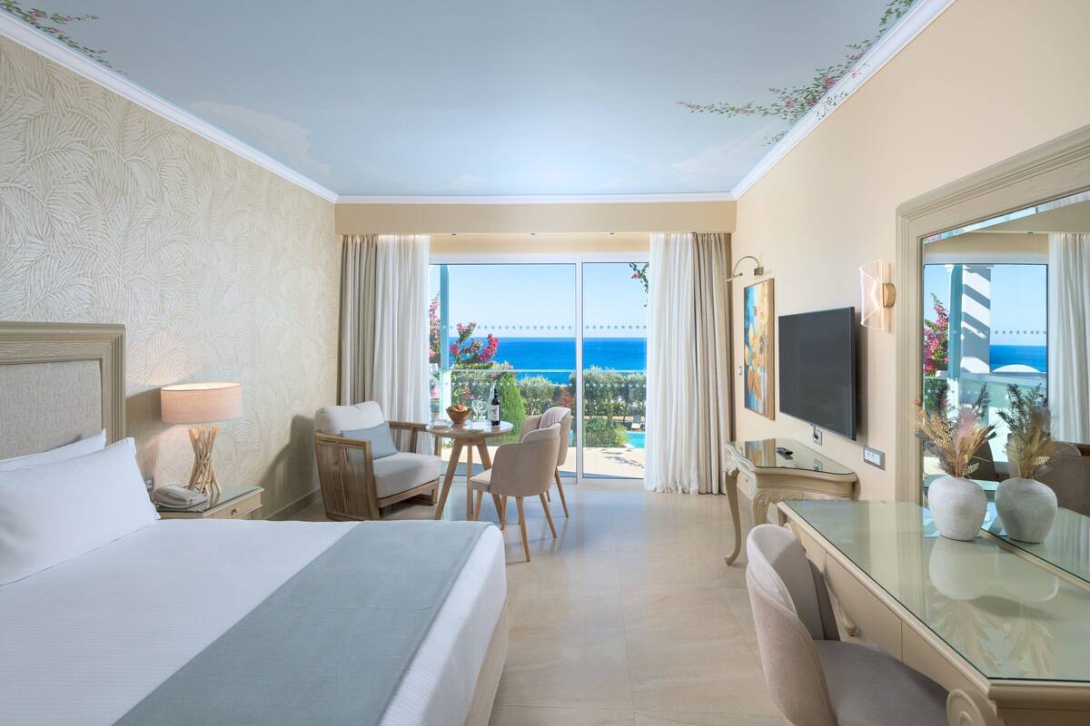 Hotelkamer met een bed, bureau, stoel, televisie en een groot raam met uitzicht op de oceaan.