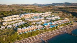 Vista aérea de un complejo turístico con varias edificaciones, una playa con filas de tumbonas y un amplio espacio para piscina.