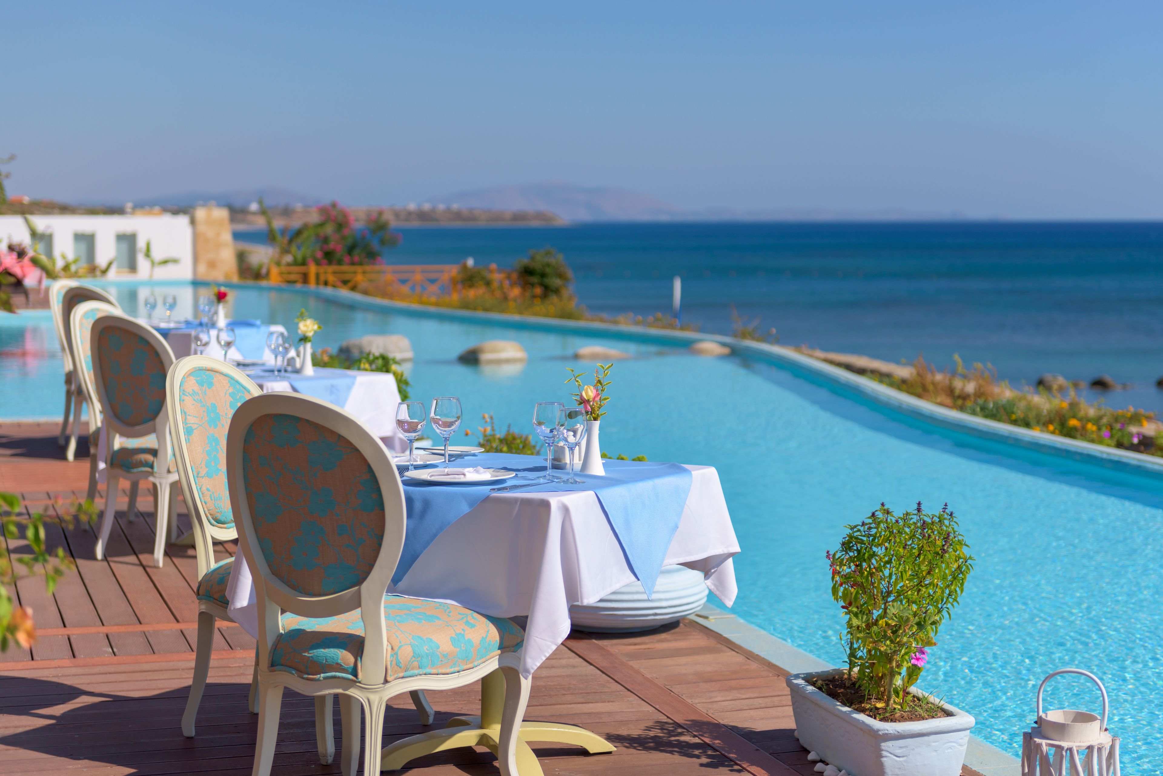 A dining area with tables and chairs by a pool, overlooking the sea.