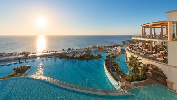 Un resort con una amplia piscina infinita, vista al océano iluminado por el sol y una zona de estar al aire libre techada.
