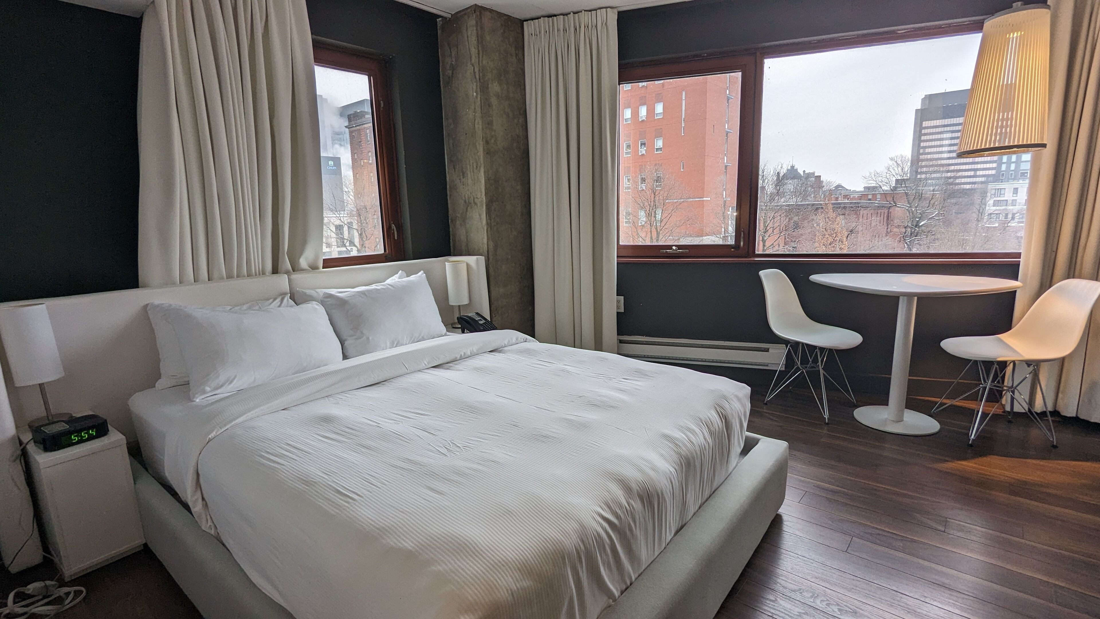 A hotel room with a bed, bedside table, lamp, and a view of the city through large windows.
