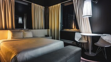 A hotel room with a bed, bedside table, chair, and a desk with a lamp.