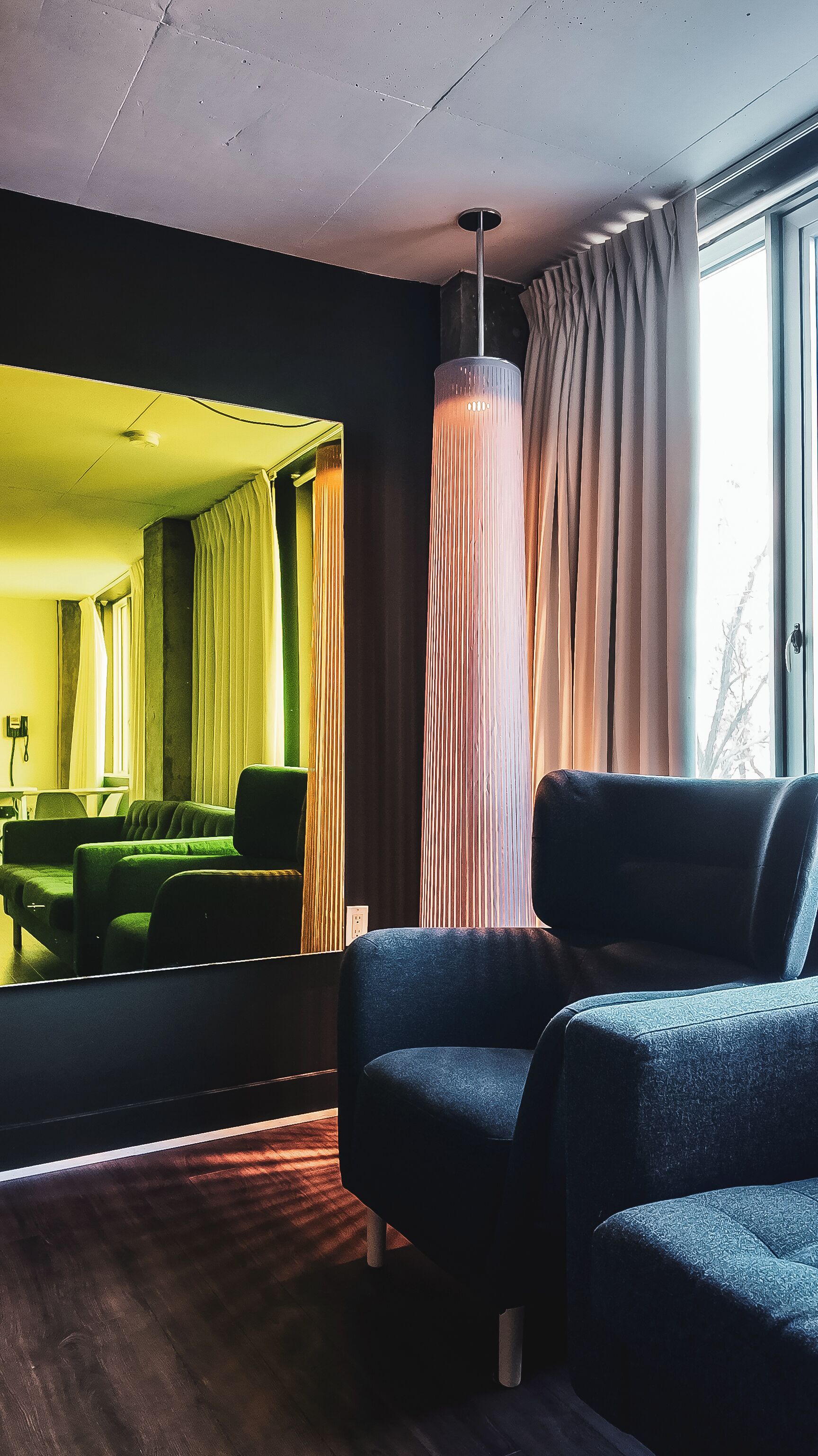 Una habitación moderna con un espejo grande, un sofá verde, un sillón azul y una lámpara alta y cilíndrica.