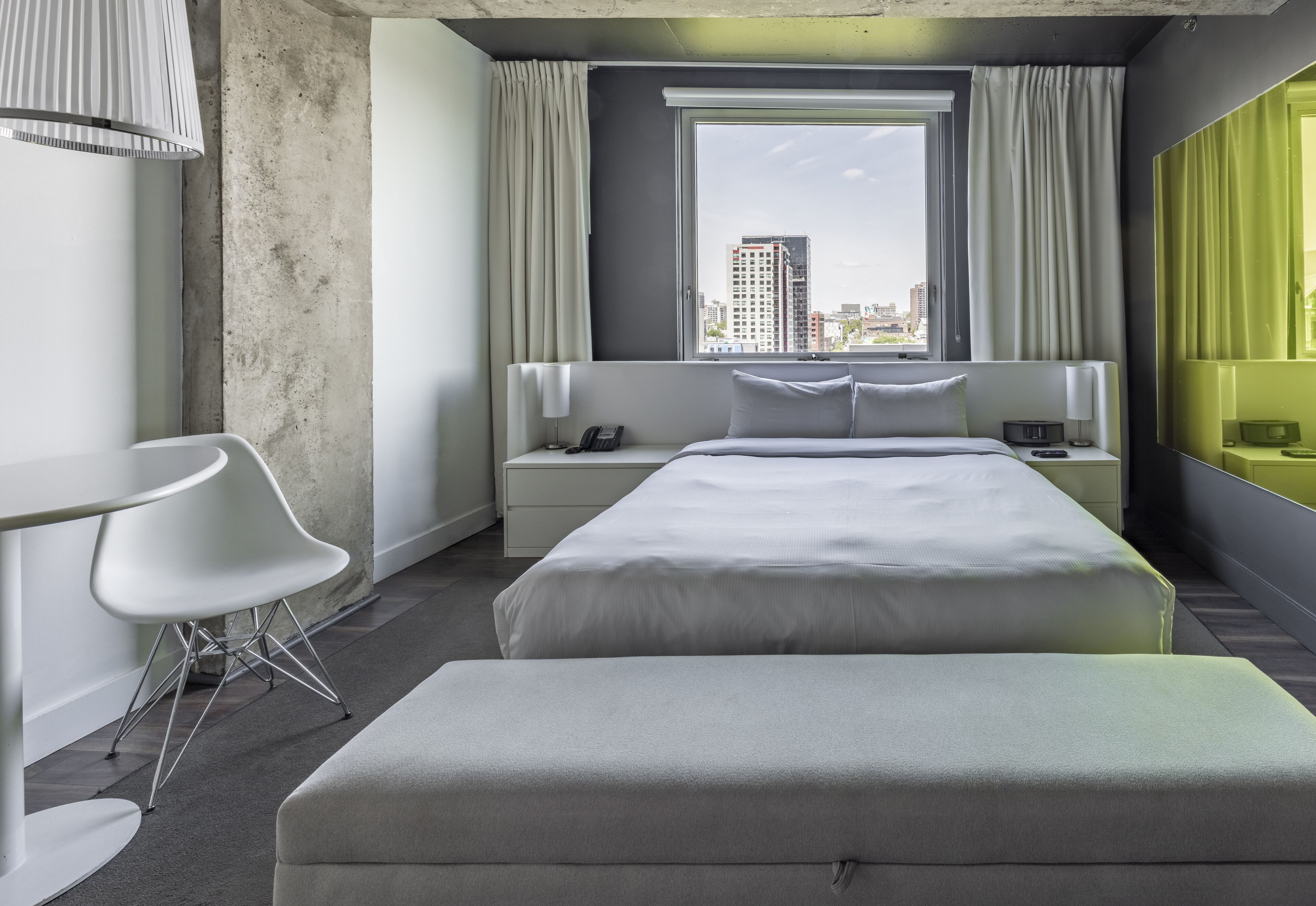 A modern hotel room with a large bed, a bedside table with a lamp, a chair, and a view of the city through a large window.