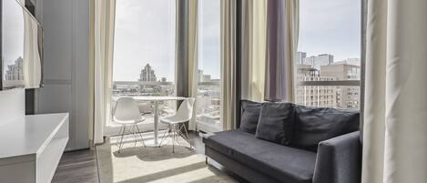 A modern living room with a grey sofa, white chairs, a small table, and a large window offering a city view.