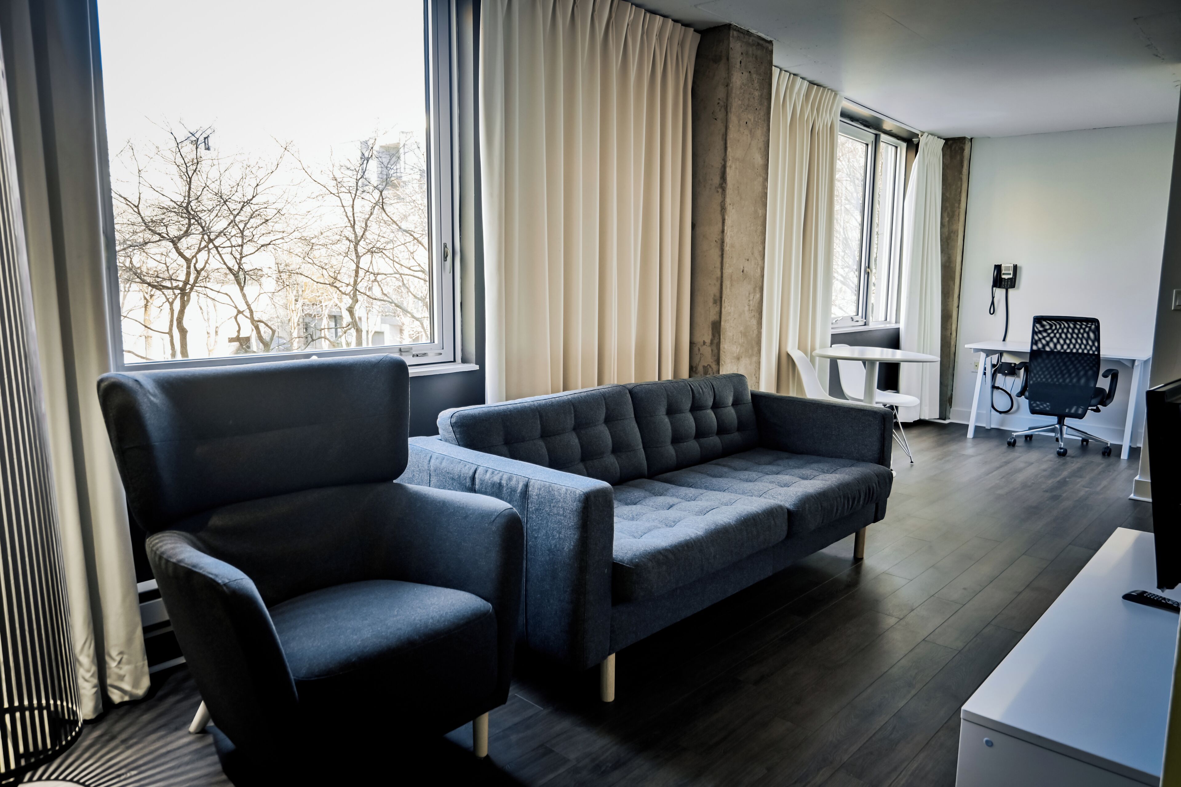A modern hotel room with a sofa, armchair, and a desk with a chair.