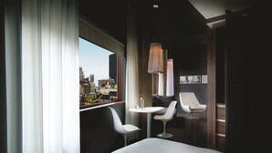Une chambre d’hôtel moderne avec un grand lit, deux chaises blanches, une petite table et une vue sur la ville par une grande fenêtre.
