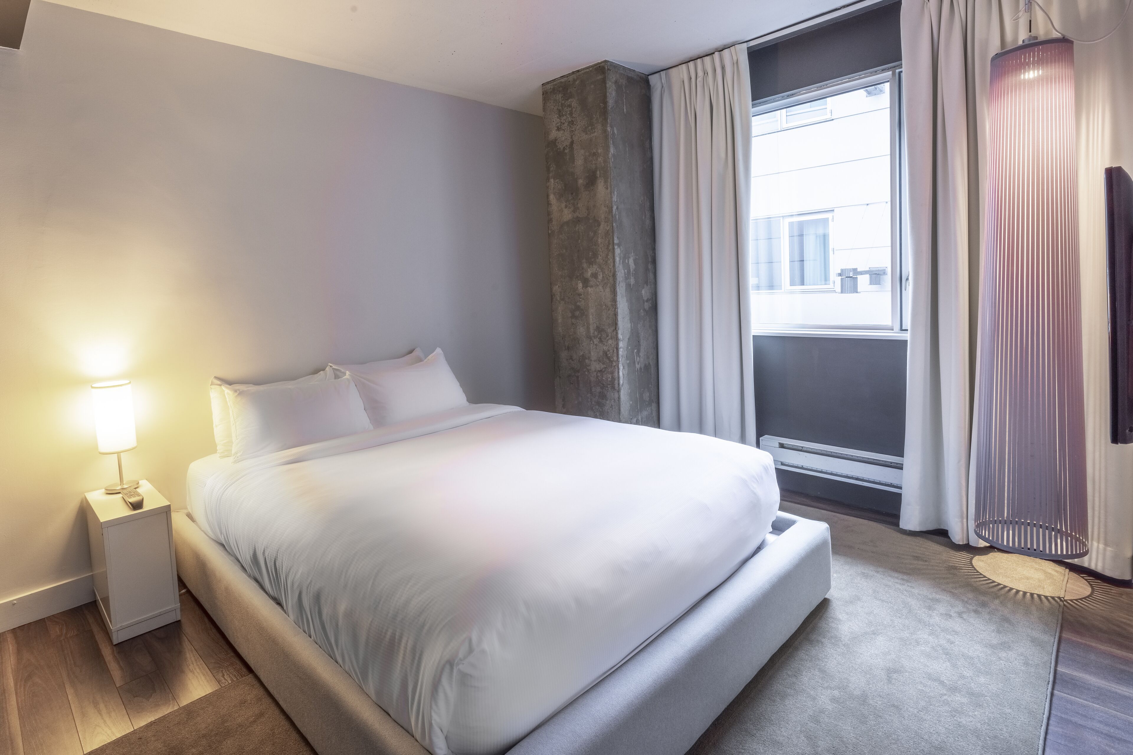 Una habitación de hotel moderna con una cama grande, una mesita de noche con lámpara, un ventanal con cortinas y una lámpara de pie.