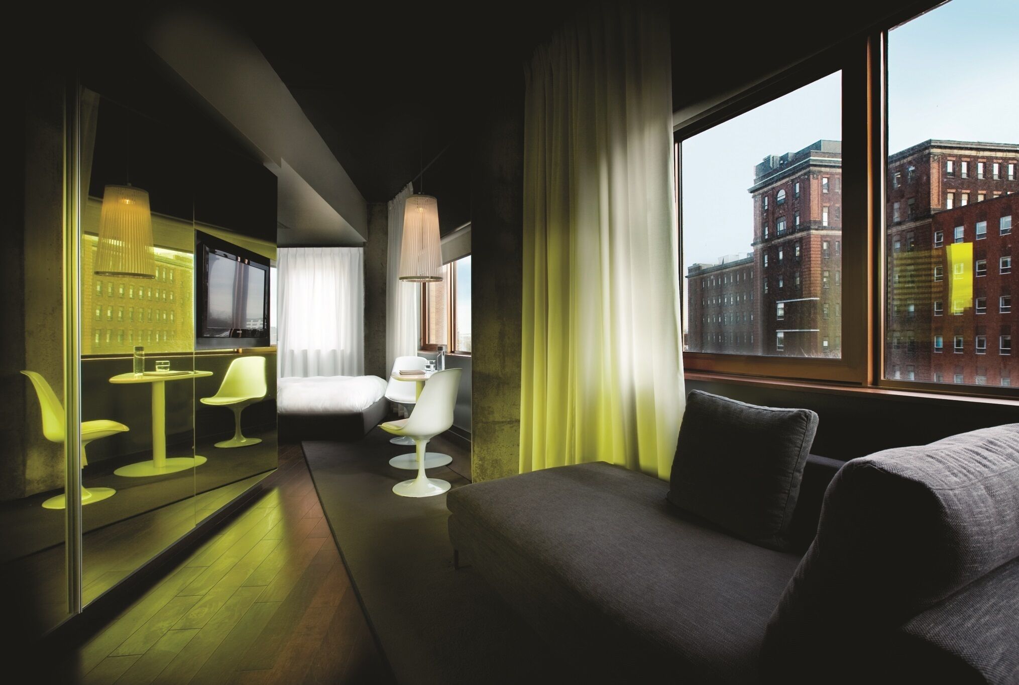 Una habitación de hotel moderna con sofá, zona de comedor con sillas, televisor y ventana grande con vista a la ciudad.