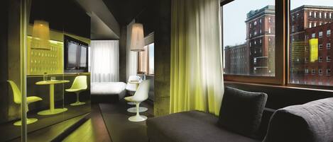 A modern hotel room with a sofa, a dining area with chairs, a TV, and a large window with a city view.