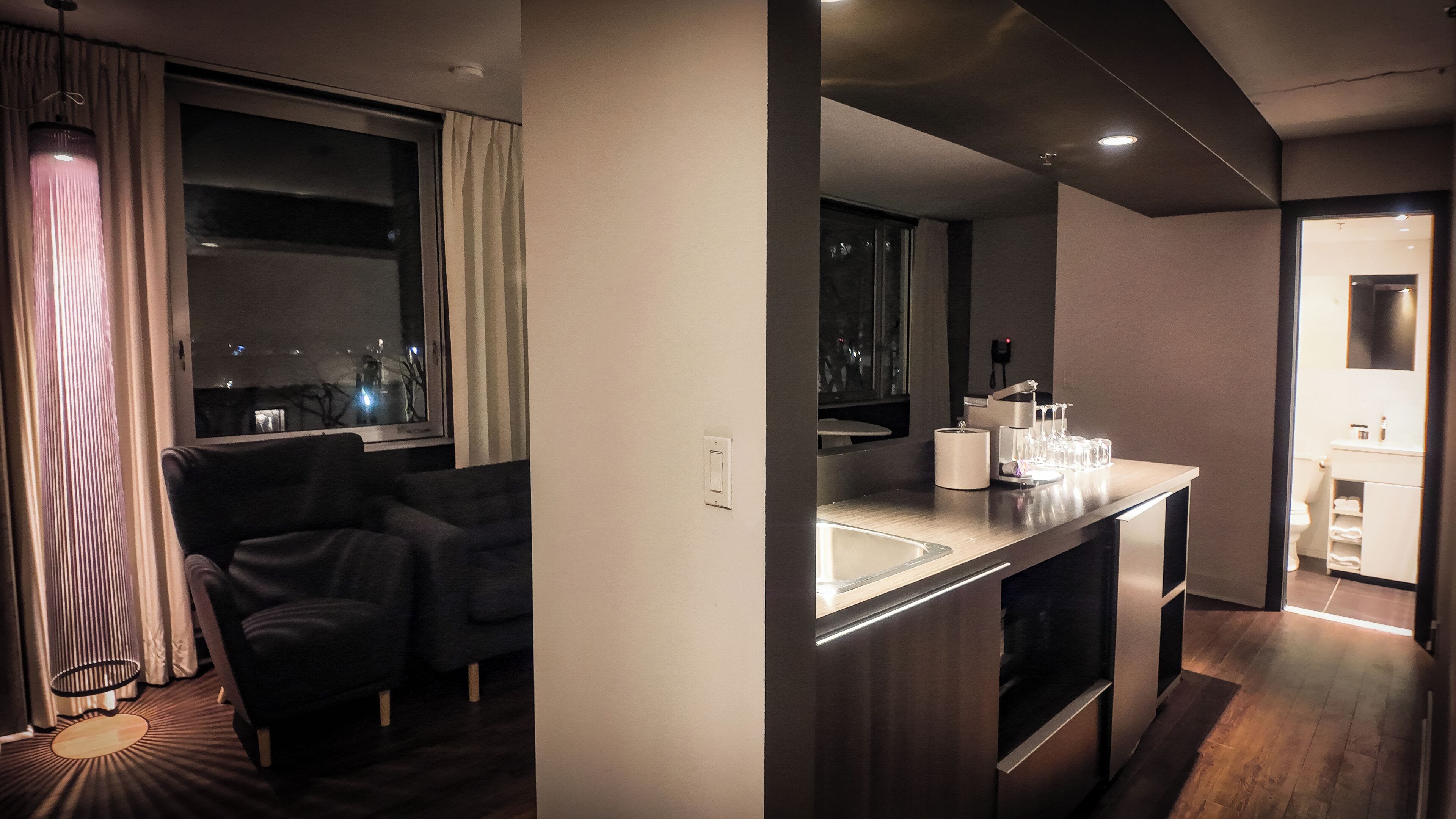 A modern hotel room with a sofa, a dining area with a bar, and a view of the city at night.