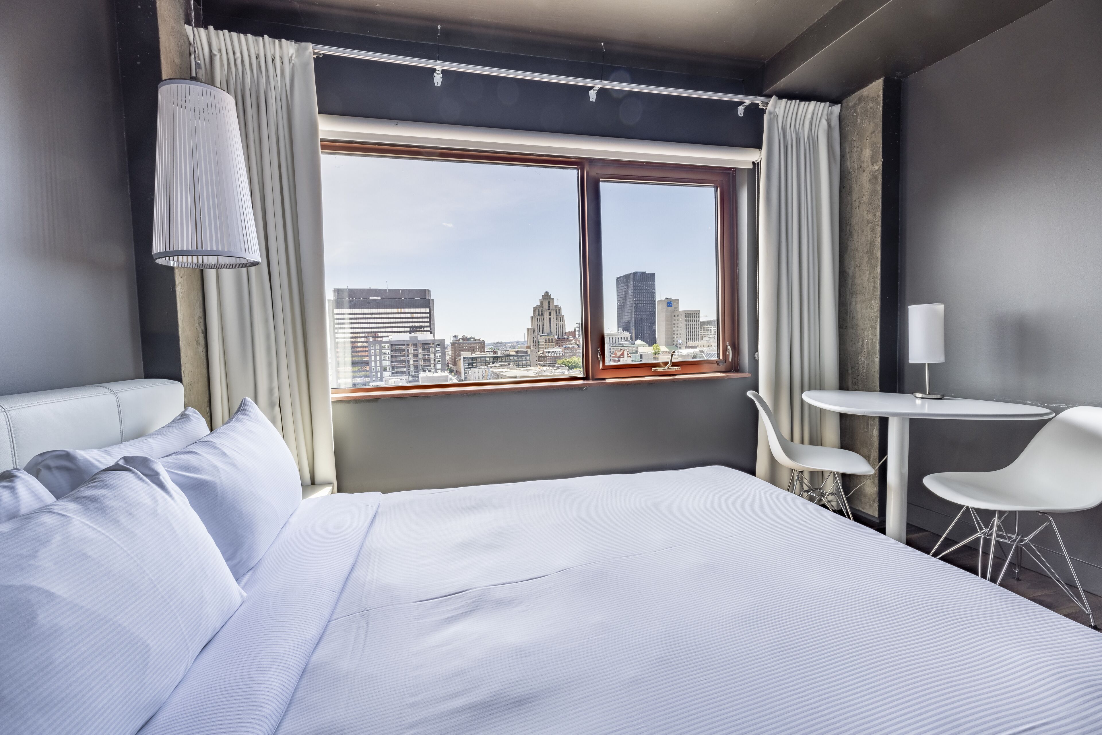 Habitación de hotel moderna con una cama grande, una mesita y vistas a la silueta urbana a través de un ventanal amplio.