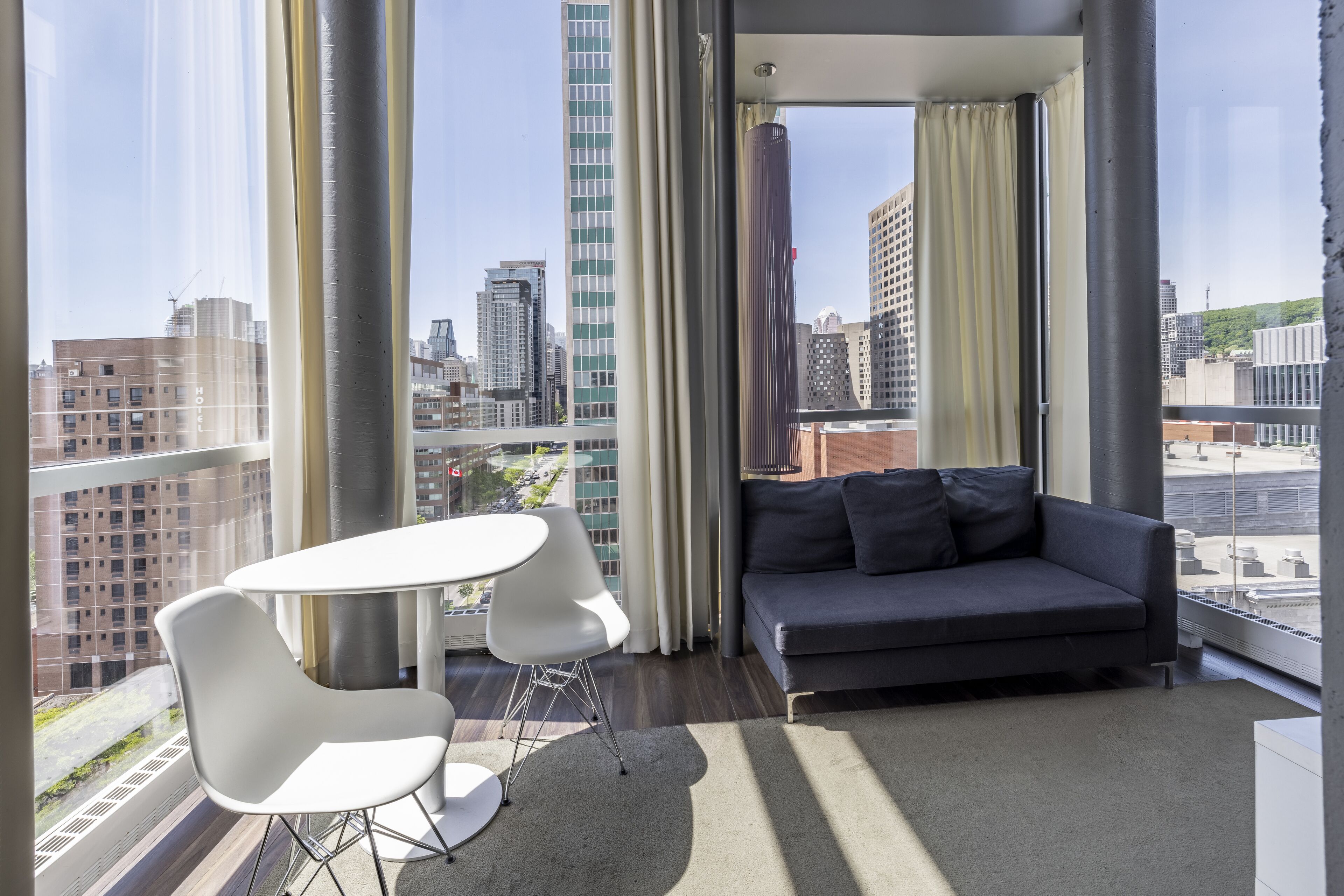 A modern living room with a sofa, a small table, and two chairs, large windows with curtains, and a city view.