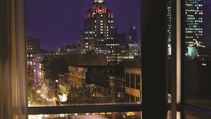 Une chambre moderne offrant une vue sur le paysage urbain nocturne, avec un fauteuil blanc et une petite table sur laquelle repose un magazine.