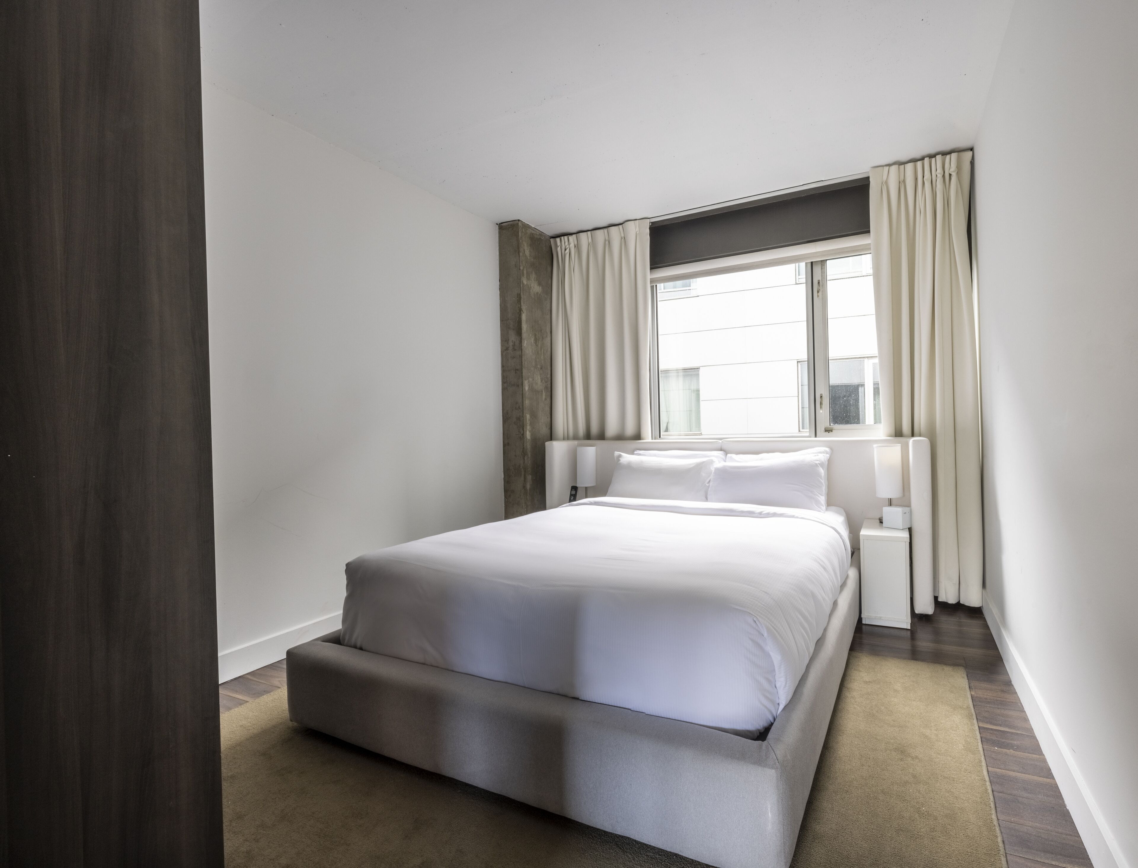 A modern hotel room with a large bed, a nightstand, a window with curtains, and a view of the city.