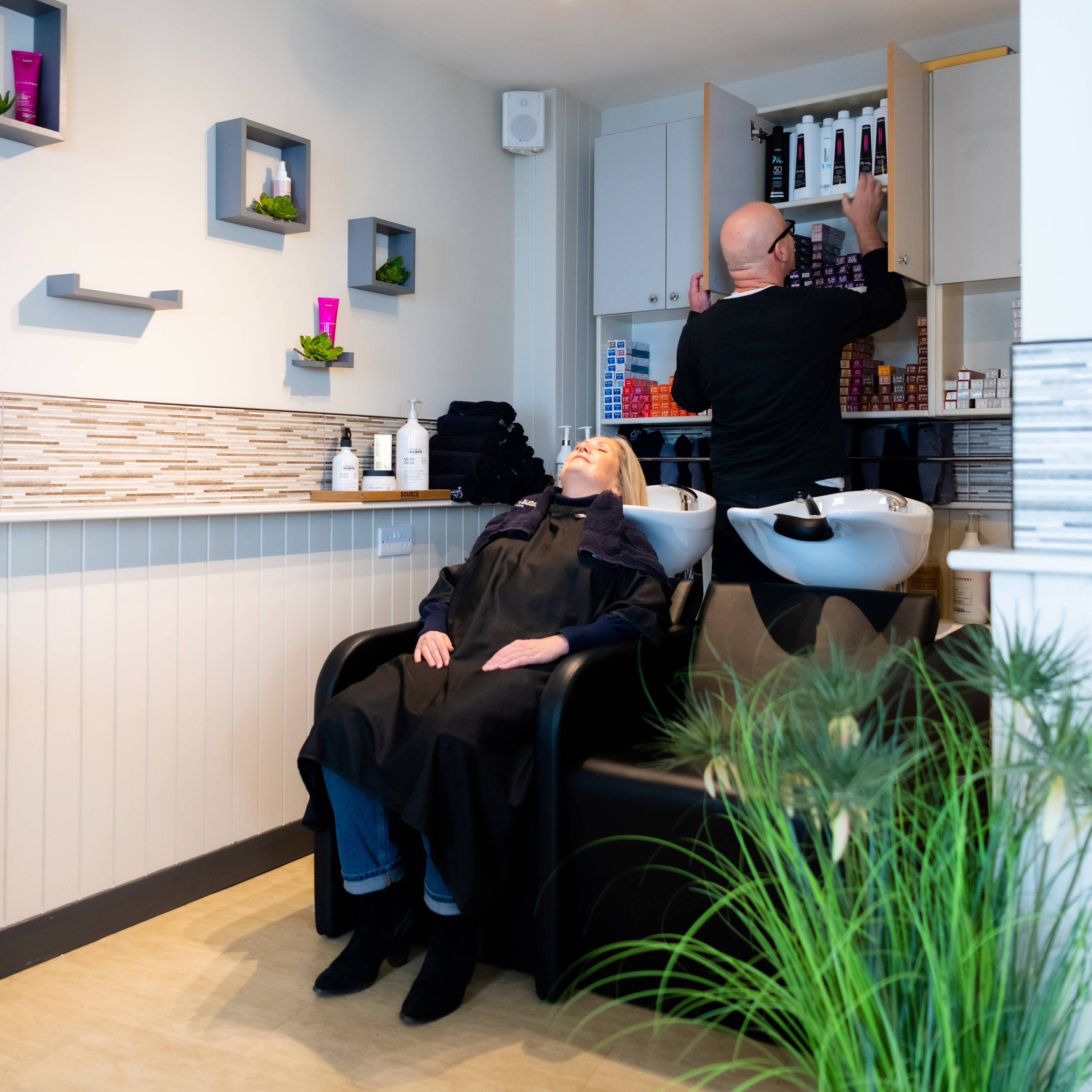 A salon with a client seated in a chair, a stylist working on the client, and shelves stocked with hair care products.
