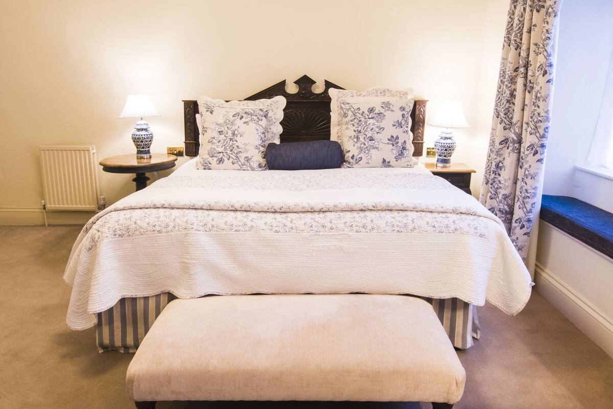 Un lit bien fait, recouvert d’une couvre-lit à motifs floraux, avec deux oreillers assortis et un banc au pied du lit. On y trouve deux tables de chevet avec des lampes, un radiateur et une fenêtre avec des rideaux.