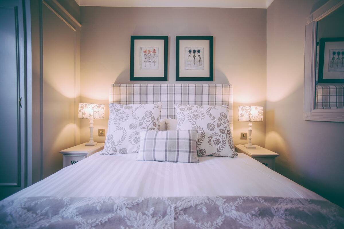 Un lit bien fait, agrémenté d’oreillers à motifs floraux et d’un plaid à carreaux, avec deux tables de chevet de chaque côté, sur lesquelles sont posées des lampes.