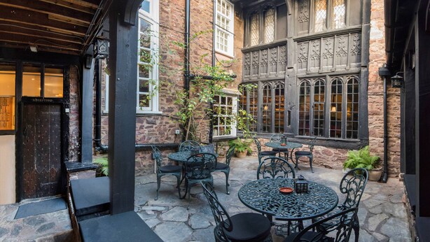 A courtyard with stone flooring, metal tables and chairs, and a brick building with large windows. - Luttrell Arms Hotel (Minehead)