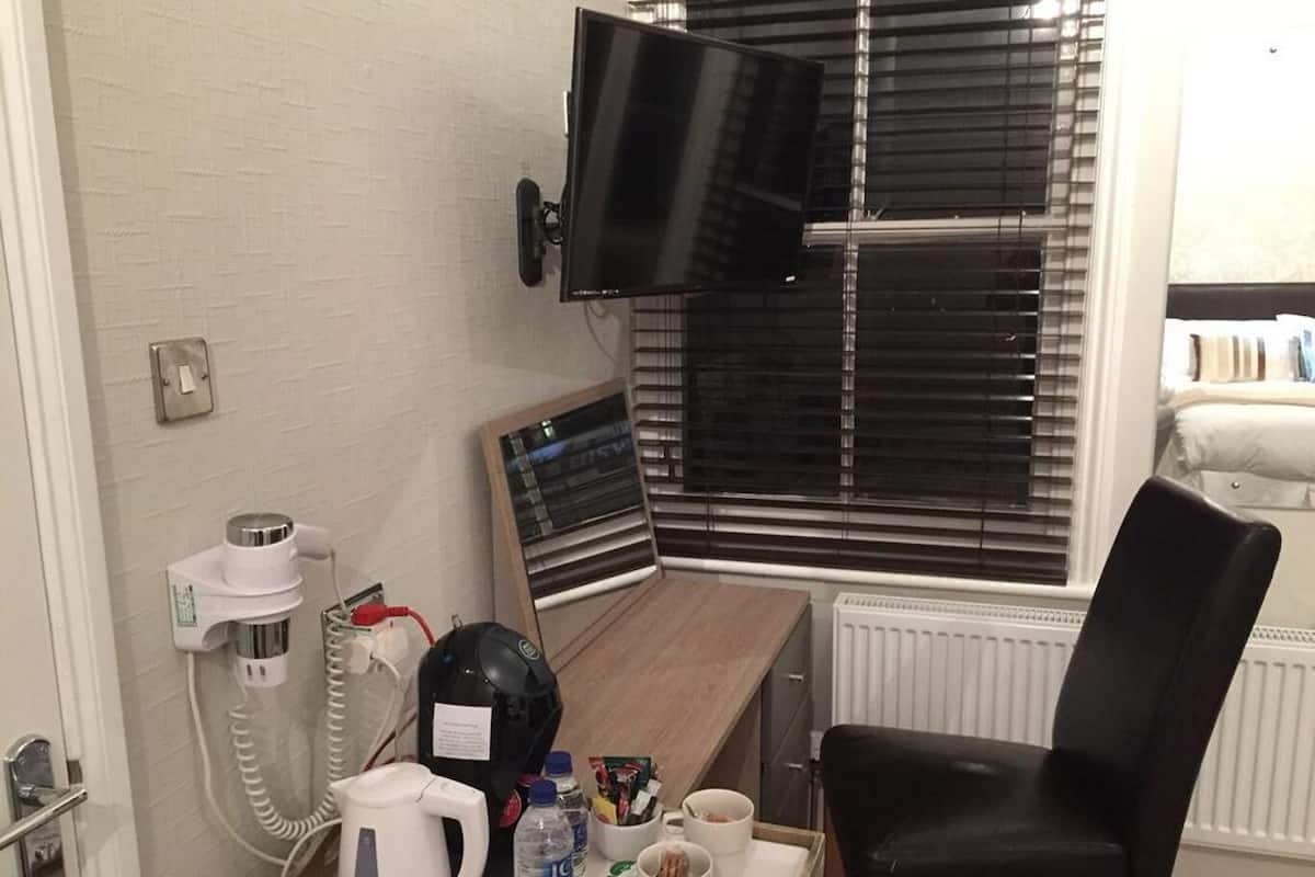 A hotel room with a bed, a TV mounted on the wall, a coffee maker, a kettle, a tray with snacks and drinks, and a window with blinds.