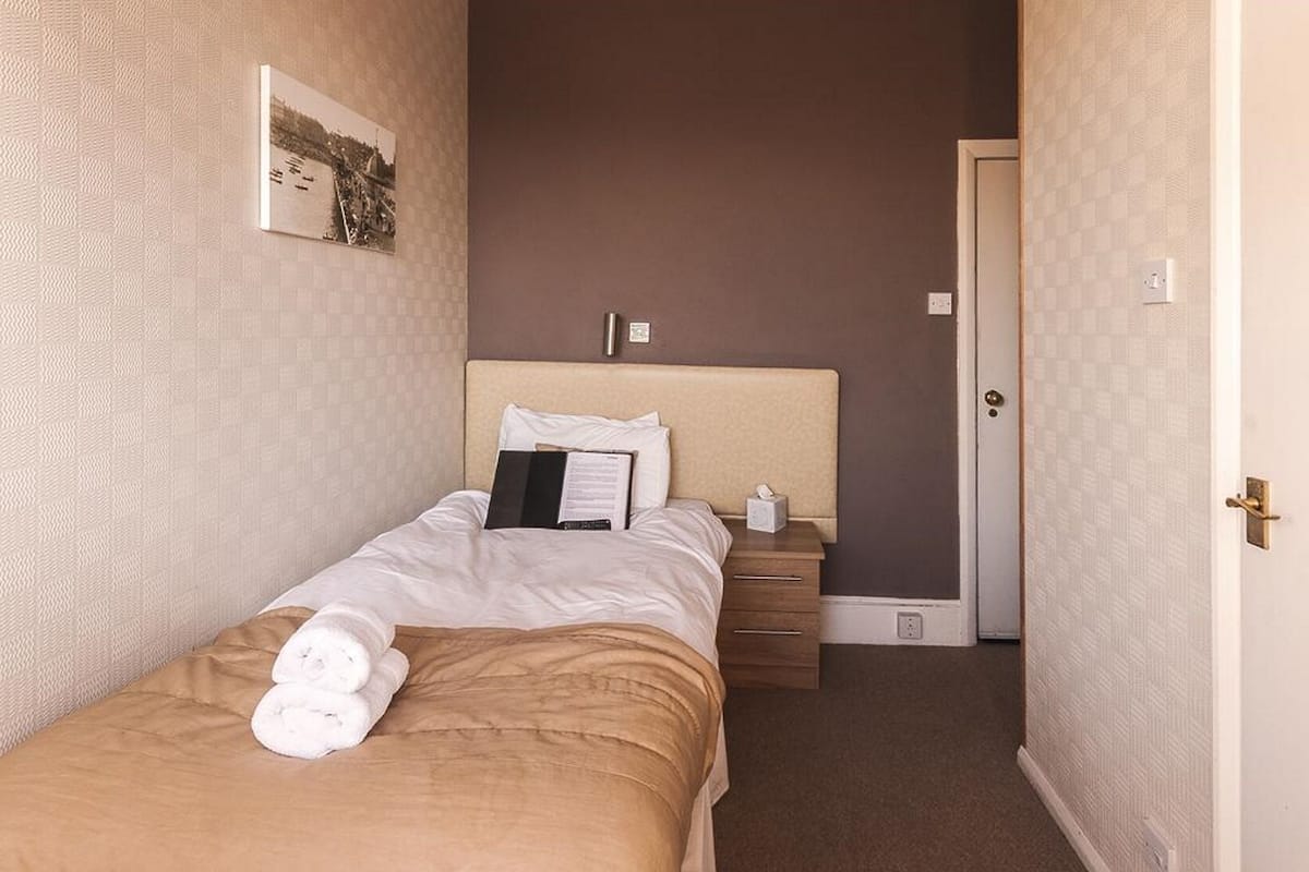 A hotel room with a single bed, a bedside table, a nightstand, a door, and a wall-mounted picture.