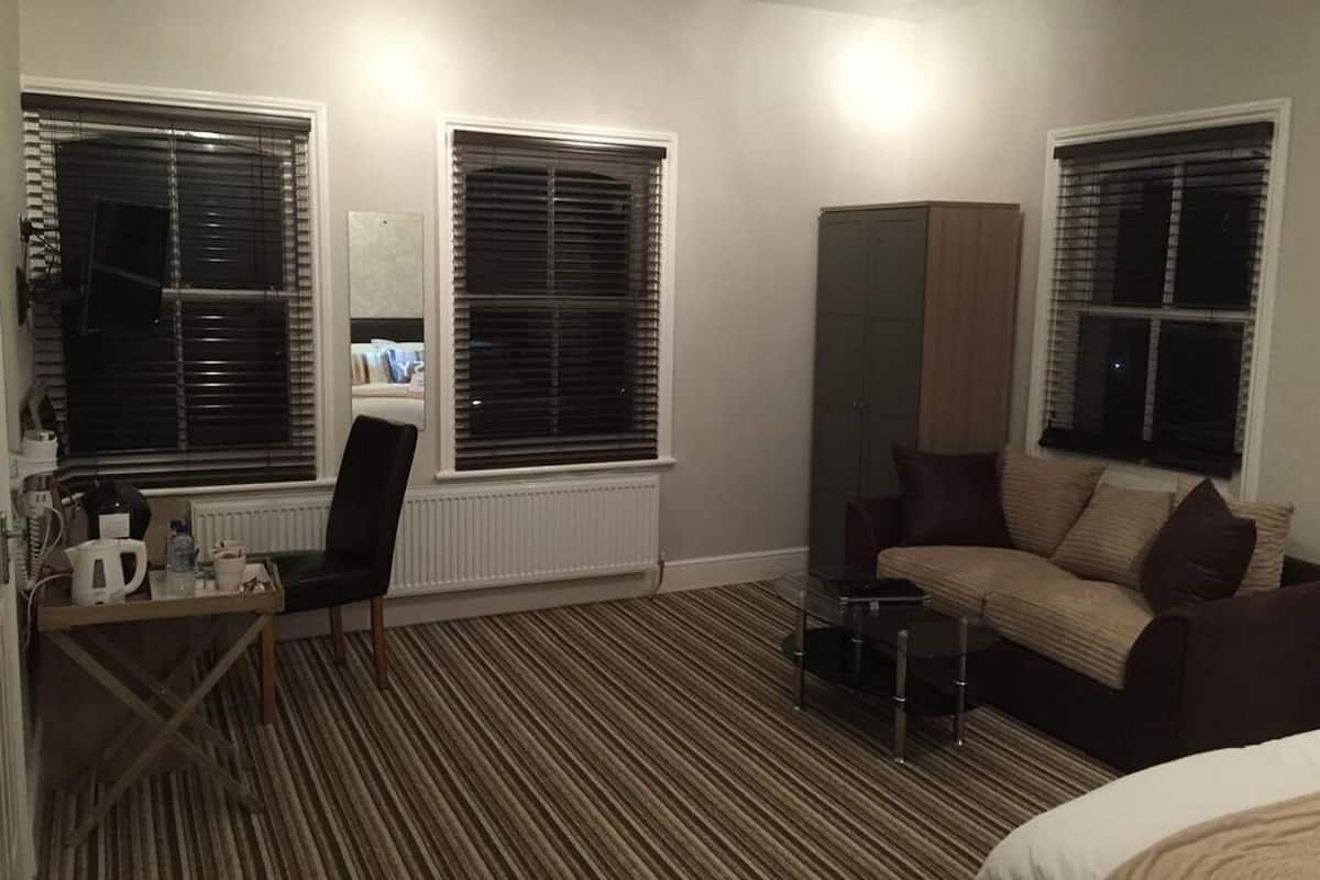 A hotel room with a striped carpet, a sofa, a small table, a cabinet, and two windows with blinds.