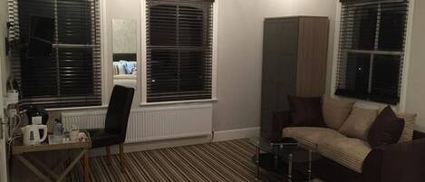 A hotel room with a striped carpet, a sofa, a small table, a cabinet, and two windows with blinds.