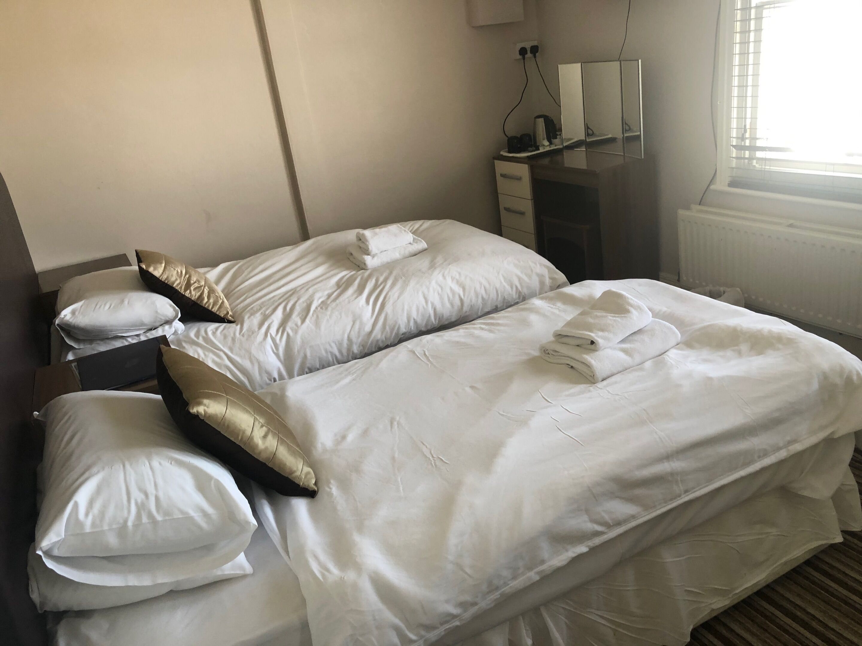 A neatly made bed with white linens and a few pillows, a small bedside table with a lamp, and a window with blinds.