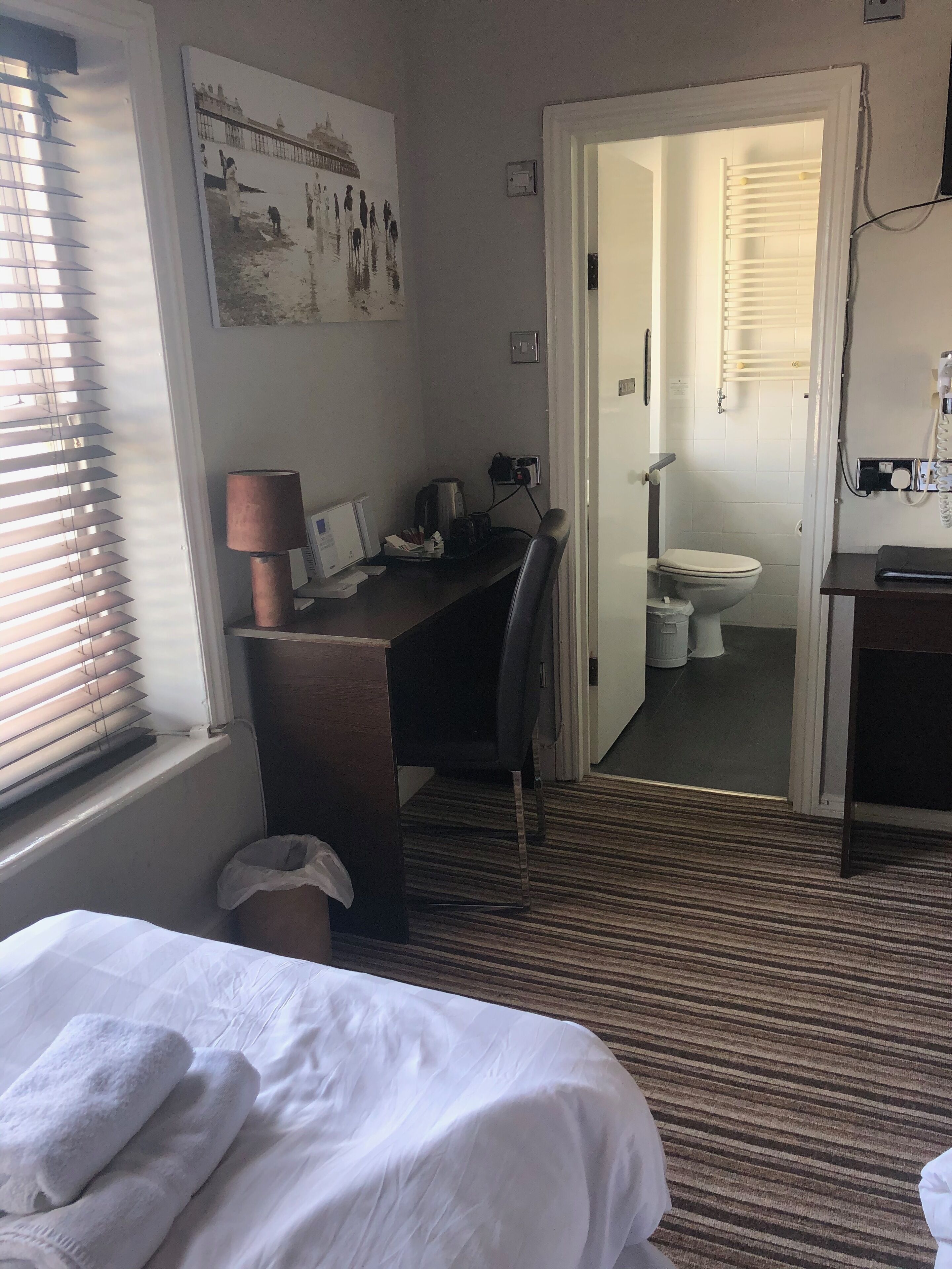 Une chambre d’hôtel avec un lit, un bureau avec un ordinateur, une chaise, une lampe et une salle de bain visible à travers une porte ouverte.
