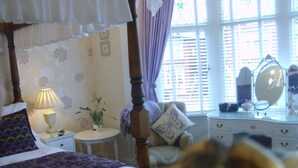 Un dormitorio con cama con dosel, una mesita de noche con lámpara, una pequeña mesa redonda con planta, una silla con cojín de mariposa y un tocador con espejo.