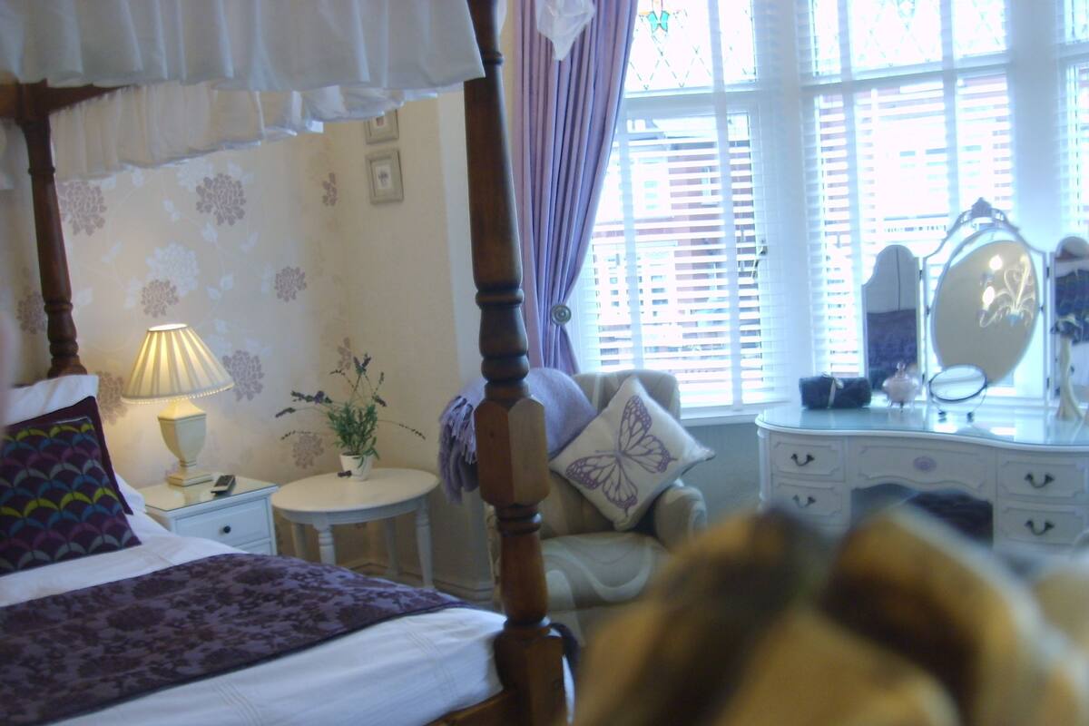 Een slaapkamer met een hemelbed, een nachtkastje met een lamp, een klein rond tafeltje met een plant, een stoel met een kussen in de vorm van een vlinder en een kaptafel met een spiegel.