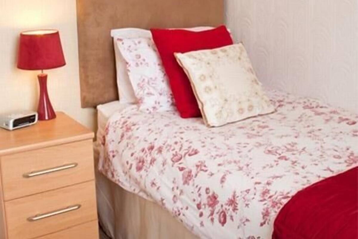 Een netjes opgemaakt bed met een bloemenbedekking, een rood kussen en een houten nachtkastje met een lamp en een klein elektronisch apparaat.