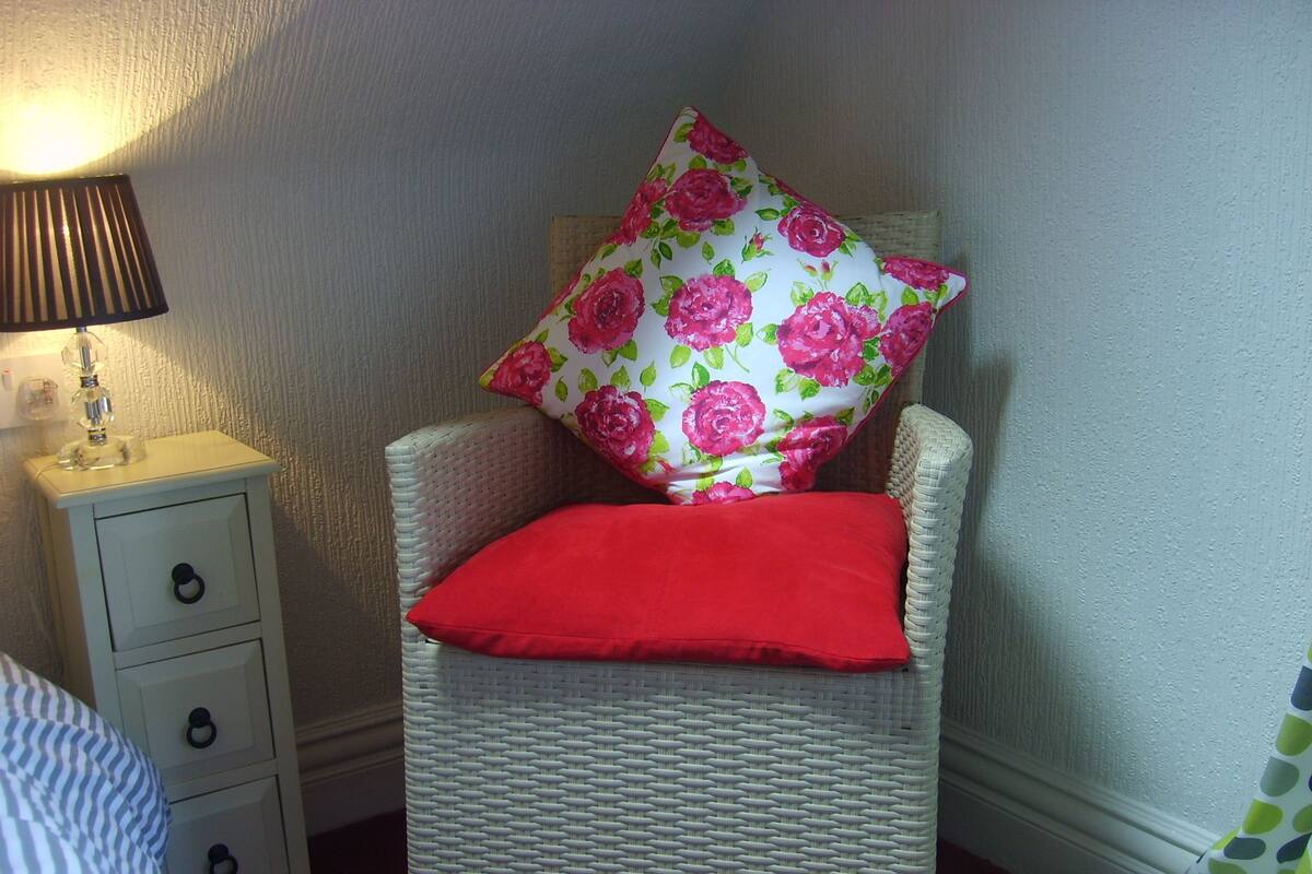Een rieten stoel met een rode kussen en een bloemenkussen, een nachtkastje met een lamp, en een witte ladekast.
