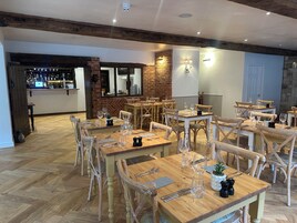 Interior de restaurante com mesas e cadeiras de madeira, parede de tijolos e área de bar ao fundo.