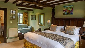 A bedroom with a large bed, wooden ceiling beams, a radiator, a window with curtains, a painting on the wall, and a sofa in the adjacent room. - The Moonraker Hotel (Trowbridge)